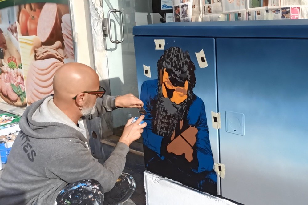 O artista Fly Pontes a aplicar retoques com spray na roupa do Remexido. O artista usa um pedaço de folha de papel para proteger manualmente áreas adjacentes (masking), garantindo a definição dos tons na roupa do guerrilheiro.