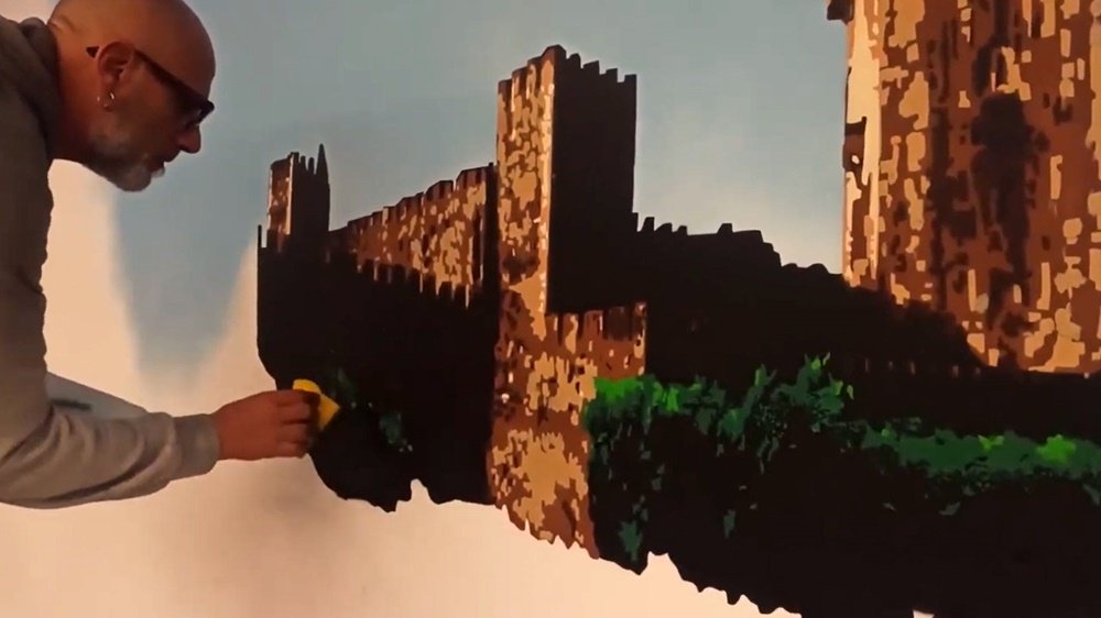 O artista Fly Pontes a aplicar os últimos retoques no Castelo de Beja com uma esponja. O Castelo está quase pronto, e o artista define os tons verdes para dar vida às árvores, complementando a aplicação de stencil no mural da BA11.