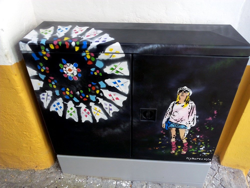 Pintura "A Rosácea da Sé" terminada num armário de eletricidade em Évora. A Rosácea estilizada brilha com cores vibrantes contra um fundo preto com salpicos. Uma figura de turista admira a obra, prestando homenagem ao património de Évora.