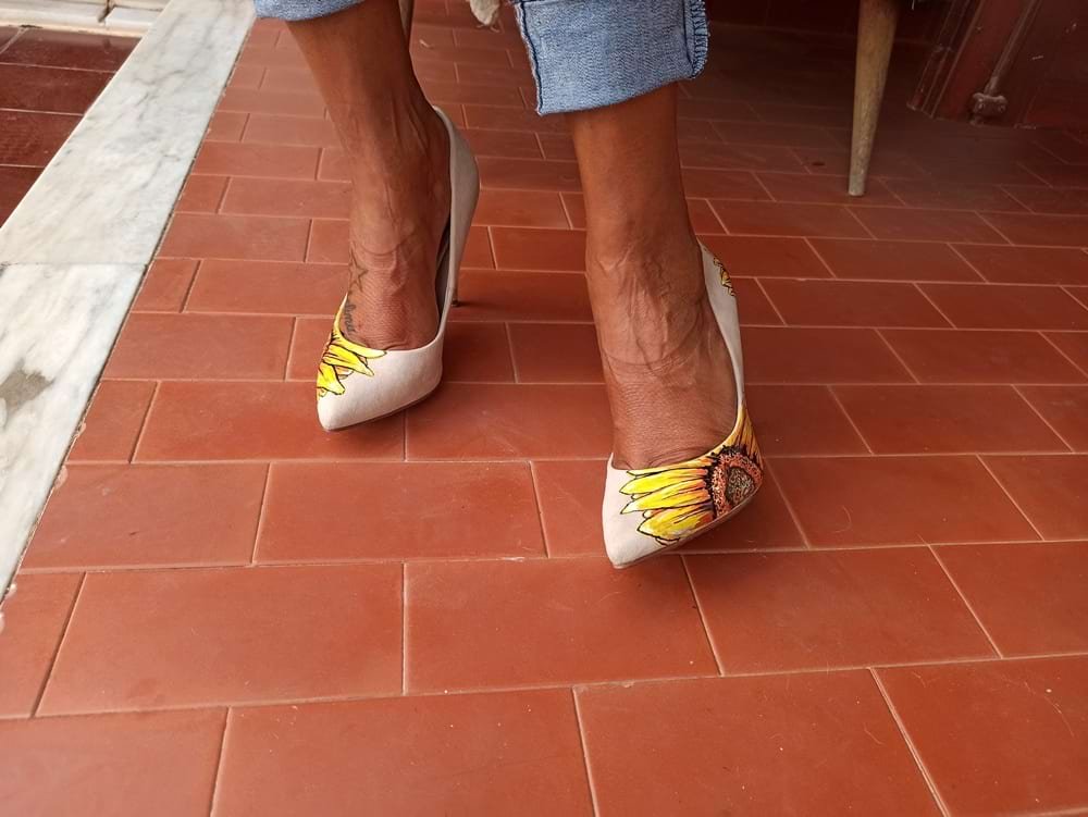Sapatos stiletto de salto alto personalizados com o design de um Girassol pintado na ponta externa. A pintura, feita com marcadores Molotow One4all, apresenta tons vibrantes de amarelo e laranja, contrastando com o material sóbrio do calçado.