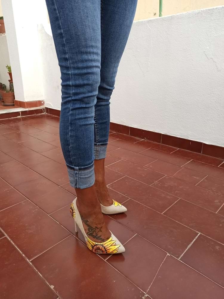 Sapatos stiletto de salto alto personalizados com o design de um Girassol pintado na ponta externa. A pintura, feita com marcadores Molotow One4all, apresenta tons vibrantes de amarelo e laranja, contrastando com o material sóbrio do calçado.