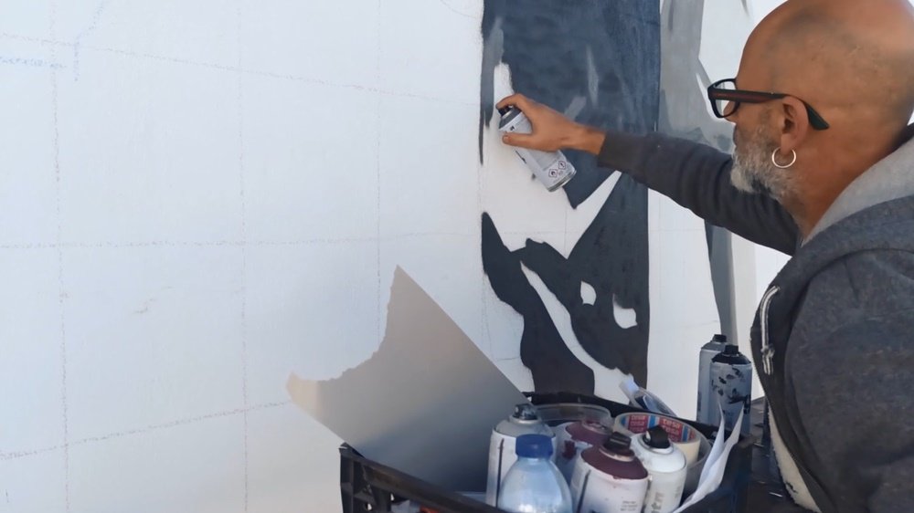 Close-up do artista Fly Pontes a pintar uma parte do mural "A Memória da Estação de Comboios" na Estação Almancil - Neche (Esteval). Vê-se uma caixa com latas de spray de diversas cores. Foco no processo de criação da arte urbana.