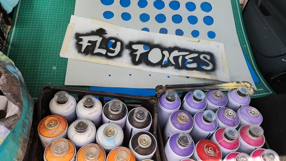 Latas de spray em tons de violeta, rosa, laranja e amarelo para o mural Pop Art da Escola Secundária Manuel Teixeira Gomes. Vê-se a folha de stencil com o meu nome e o stencil para os pontos (Ben-Day Dots), inspirados em Roy Lichtenstein.