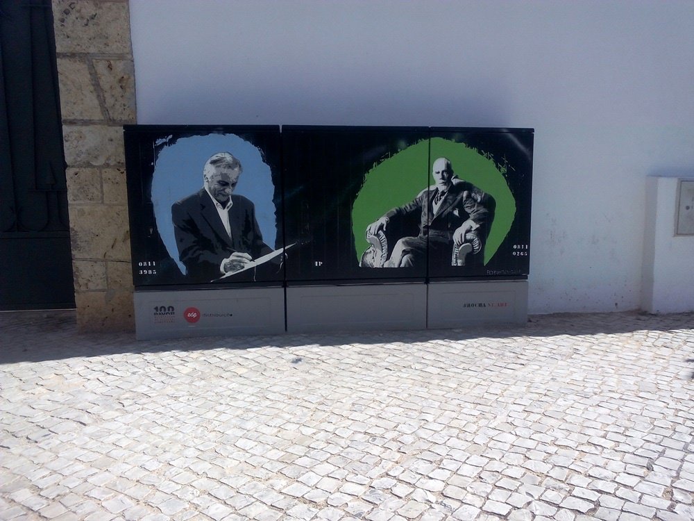 Pintura "Legados de Portimão" terminado num armário de electricidade. Apresenta Teixeira Gomes e Júlio Amaro pintados em tons de cinza, enquadrados por círculos de cor vibrante (azul e verde). Homenagem às figuras políticas e artísticas de Portimão.