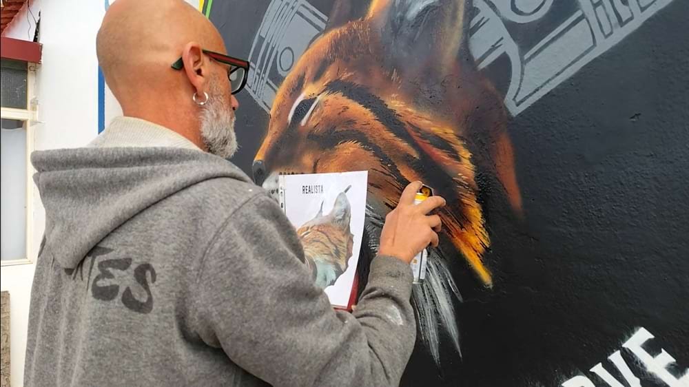 Artista a aplicar os últimos retoques no lince do mural, usando uma Tiny Pink Stencil Cap e uma foto de referência na mão.