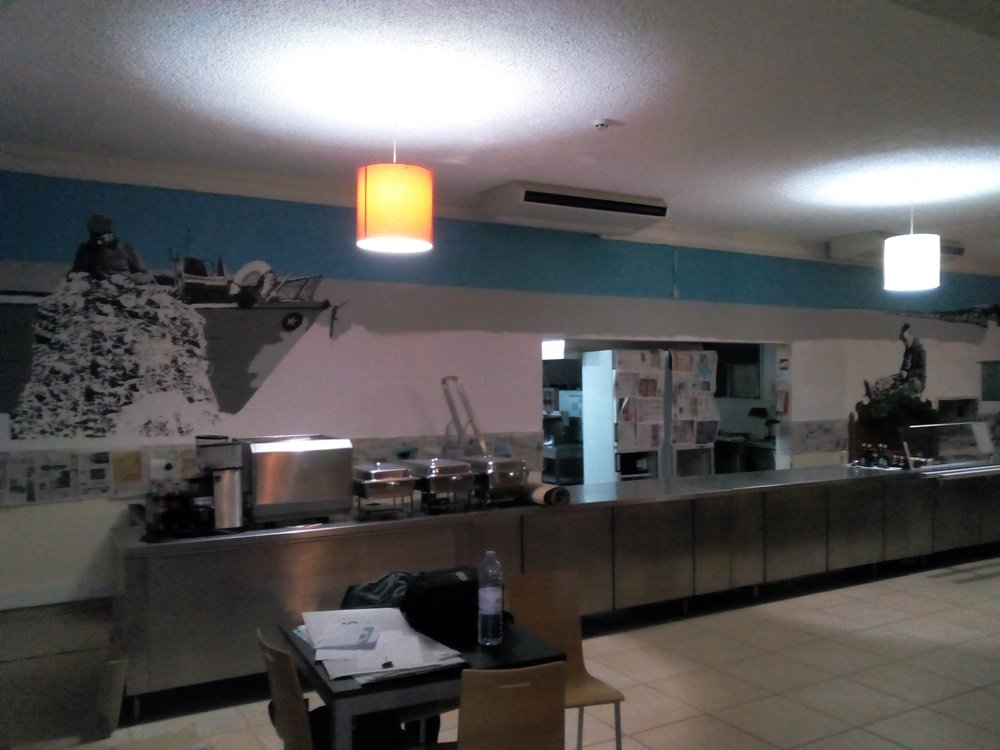 Vista afastada do mural "O Pescador de Armação de Pêra". O céu azul vibrante contrasta com os dois pescadores em tons de cinza: um a arranjar redes e outro junto a um monte. A cena de labuta marítima está na fase final, faltando apenas as gaivotas.