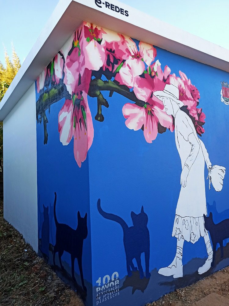 Vista que une a lateral e a frente do mural "Homenagem à Marquinhas" em Almancil. O ramo de amendoeira em flor e os gatos em silhueta ligam as faces, enquadrando a figura principal da Marquinhas em traço minimalista.