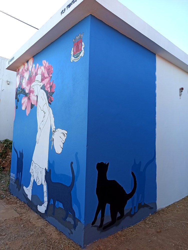 Vista que une a lateral e a frente do mural "Homenagem à Marquinhas" em Almancil. A figura minimalista da Marquinhas e o ramo de Amendoeira em Flor estão na frente, enquanto os gatos em silhueta se prolongam pela lateral, ligando as faces do PT.
