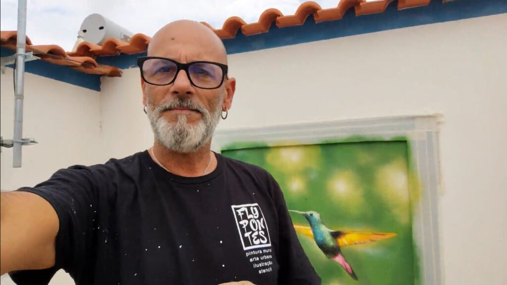 Selfie do artista Fly Pontes com mural de colibri quase terminado em fundo.