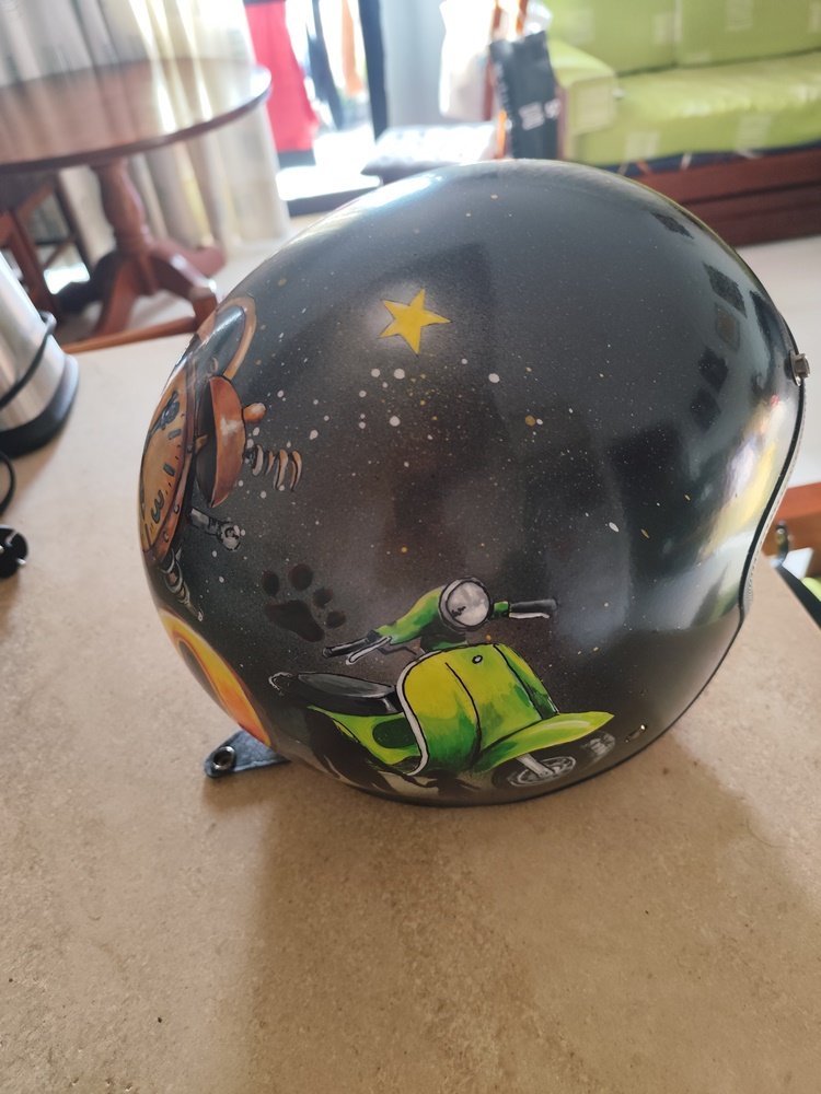 Fotografia de detalhe da parte superior de um capacete. Sobre um fundo escuro com efeitos de spray esfumado e pequenos salpicos (drops) de tinta branca, destaca-se uma pequena estrela amarela pintada com canetas de precisão. Por baixo da estrela, vê-se a parte superior de uma Vespa verde.