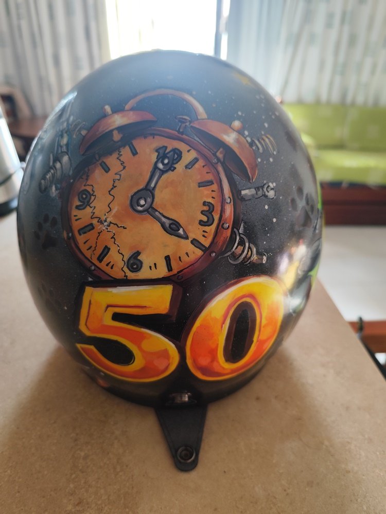 Grande plano de um capacete de motociclista terminado e envernizado com acabamento brilhante. O fundo é preto profundo, destacando a pintura manual de um número 50 e de um relógio com os ponteiros nas 04h00. A superfície reflecte a luz, mostrando a textura lisa e profissional do verniz sobre os detalhes feitos a caneta.