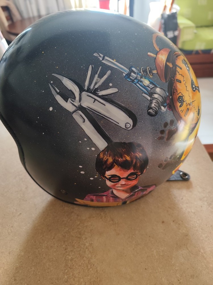 Fotografia em grande plano de um detalhe do capacete pintado à mão. Sobre um fundo escuro esfumado com spray, destacam-se as representações de um alicate e de uma chave de fendas busca-polos, pintadas com canetas de precisão, mostrando os seus detalhes metálicos. Do lado direito, vê-se parte da pintura do retrato do menino a ler.