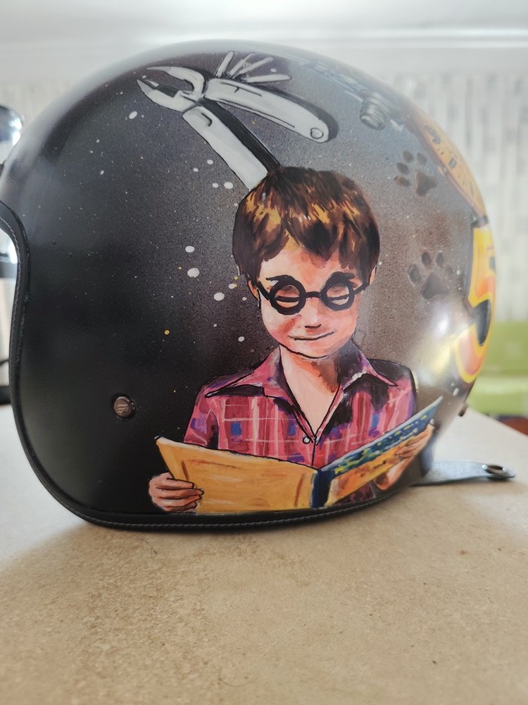 Fotografia de detalhe de um capacete pintado à mão. No centro, vê-se o retrato de uma criança pequena com óculos e sobrancelhas expressivas, simulando o acto de ler um livro. Logo acima desta imagem, está pintada a parte superior de um alicate detalhado com canetas de precisão, sobre um fundo escuro com esfumados de spray.