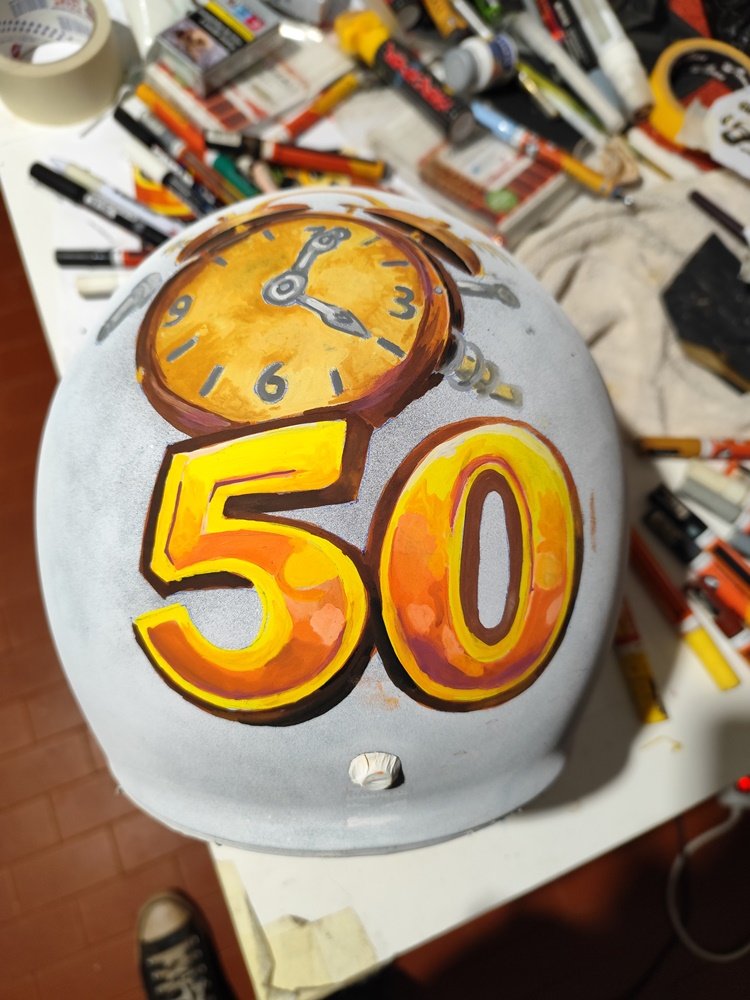 Fotografia em grande plano do processo de pintura manual num capacete branco. Sobre a superfície, vê-se o esboço do número 50 e um relógio com ponteiros. Na mesa de trabalho, estão espalhadas canetas de tinta acrílica das marcas Molotow e Posca, fitas adesivas e materiais de precisão.