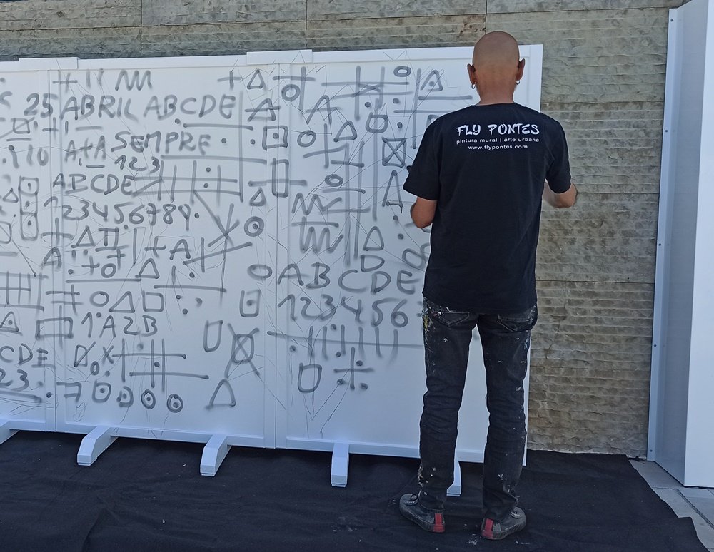 Artista Fly Pontes de costas a marcar o mural com rabiscos de referência (doodles) em painéis brancos.