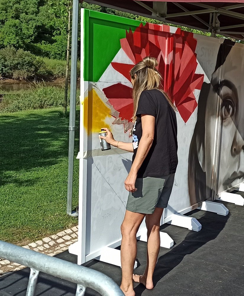 Mulher a pintar formas geométricas verdes no fundo do mural com o Rio Arade e zona verdejante ao fundo.