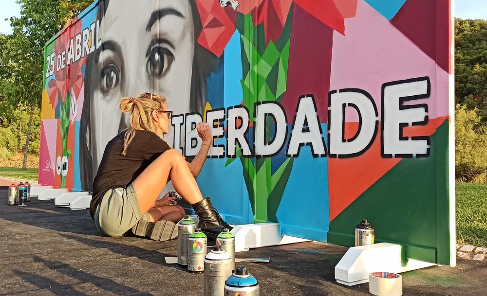 Pintura de contorno da palavra "Liberdade" com pincel, com latas de spray MTN Water Base em destaque.