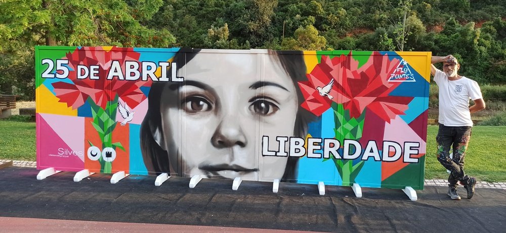 Fly Pontes encostado ao mural finalizado com o rosto da criança, cravos e pombas.