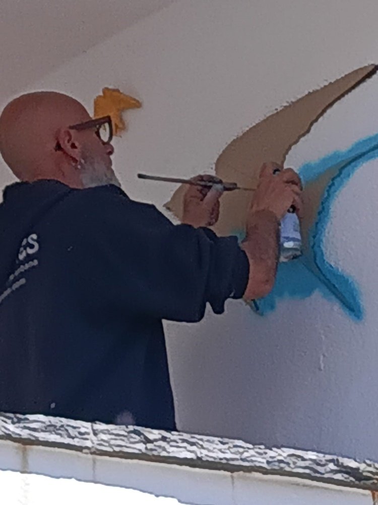artista a pintar com spray azul numa parede enquanto usa um pauzinho para segurar a máscara da pintura.