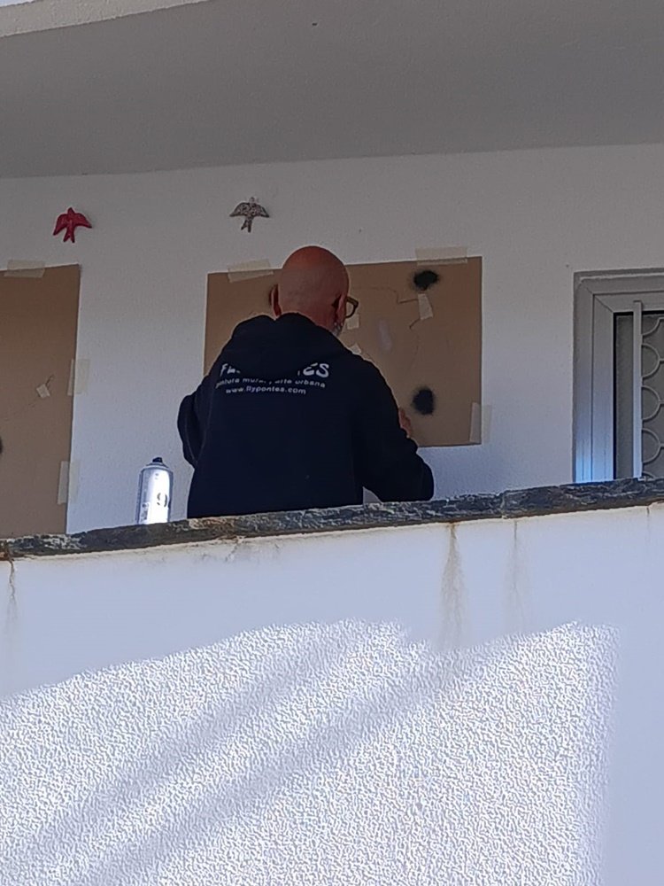 artista a aplicar spray colorido sobre um stencil multicamadas com guias de marcação visíveis na parede.