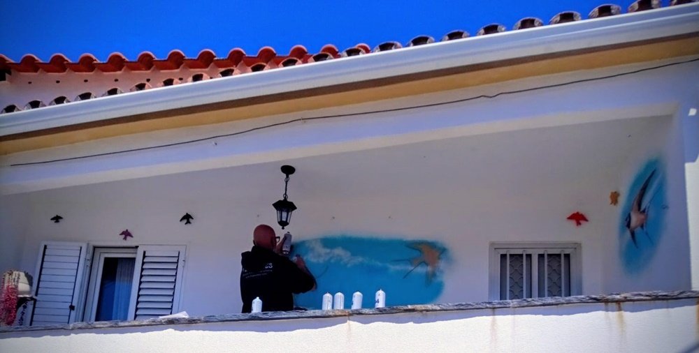 perspectiva de baixo para cima do artista a pintar o céu azul na parede de uma varanda de primeiro andar.