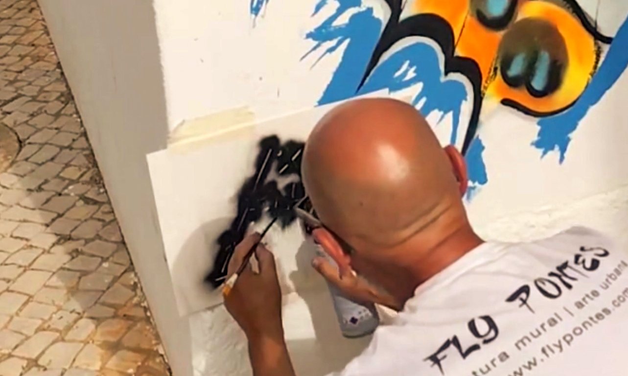 Mão do artista Fly Pontes a aplicar tinta preta a spray sobre um stencil com o logótipo oficial na parede do mural.