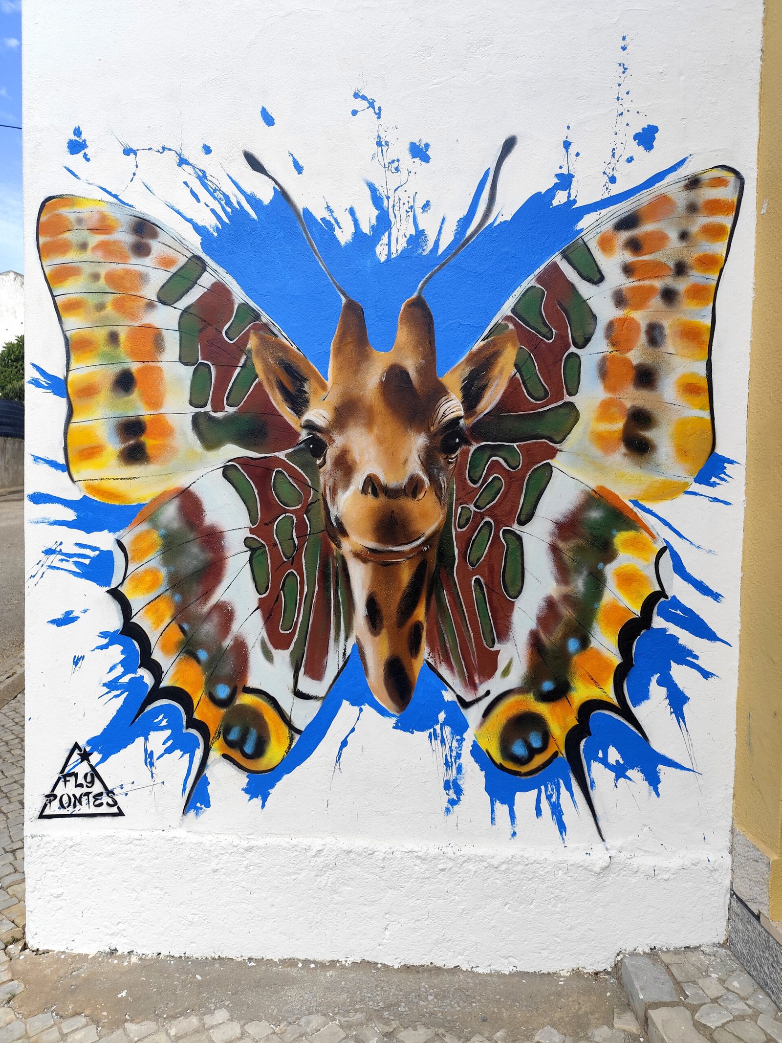 Mural finalizado de 2,5m por 3m em Messines com o rosto realista de uma girafa e asas de borboleta-do-medronheiro sobre fundo azul.