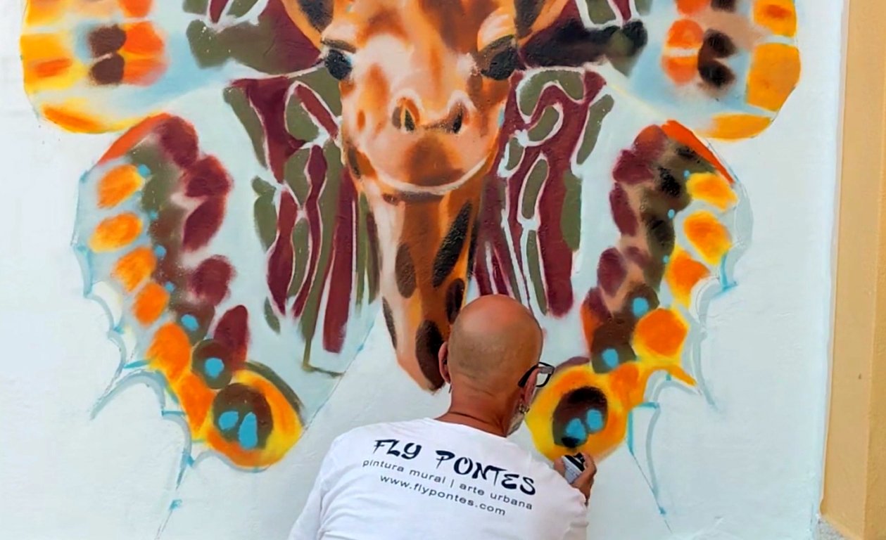 Detalhe das asas da borboleta-do-medronheiro pintadas a spray com tons de castanho, laranja e verde a nascer da nuca da girafa.